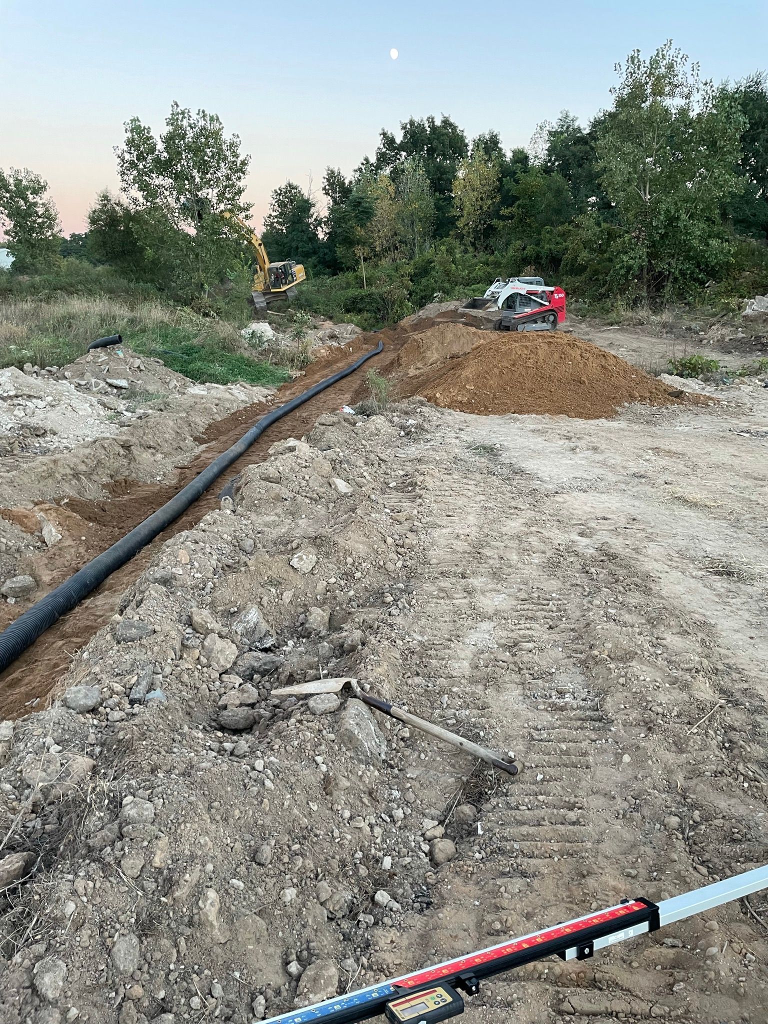 Construction site with a black pipe in a trench, dirt, trees, and equipment.