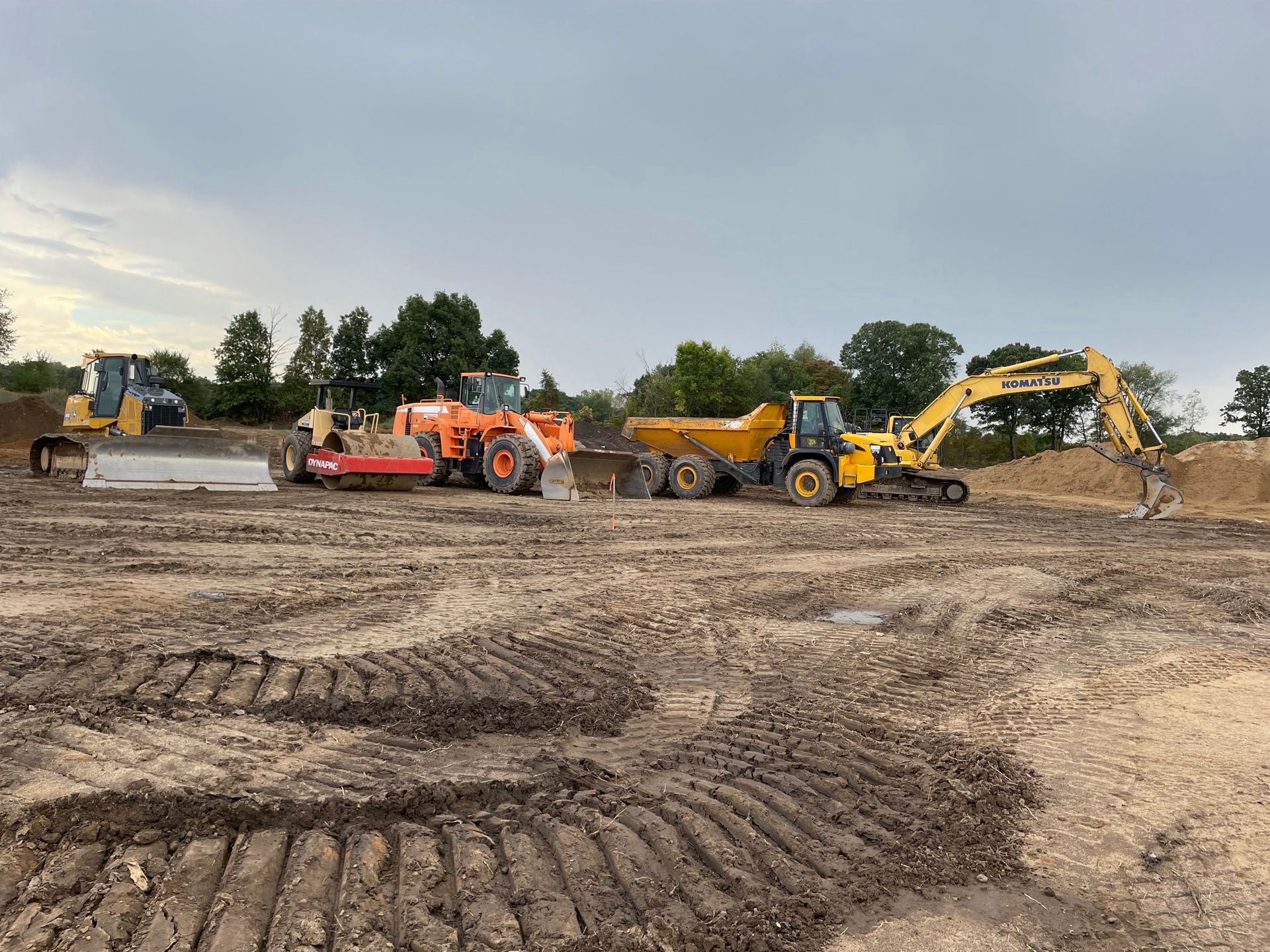 Construction site with heavy machinery including a bulldozer, roller, and excavator, on muddy ground.