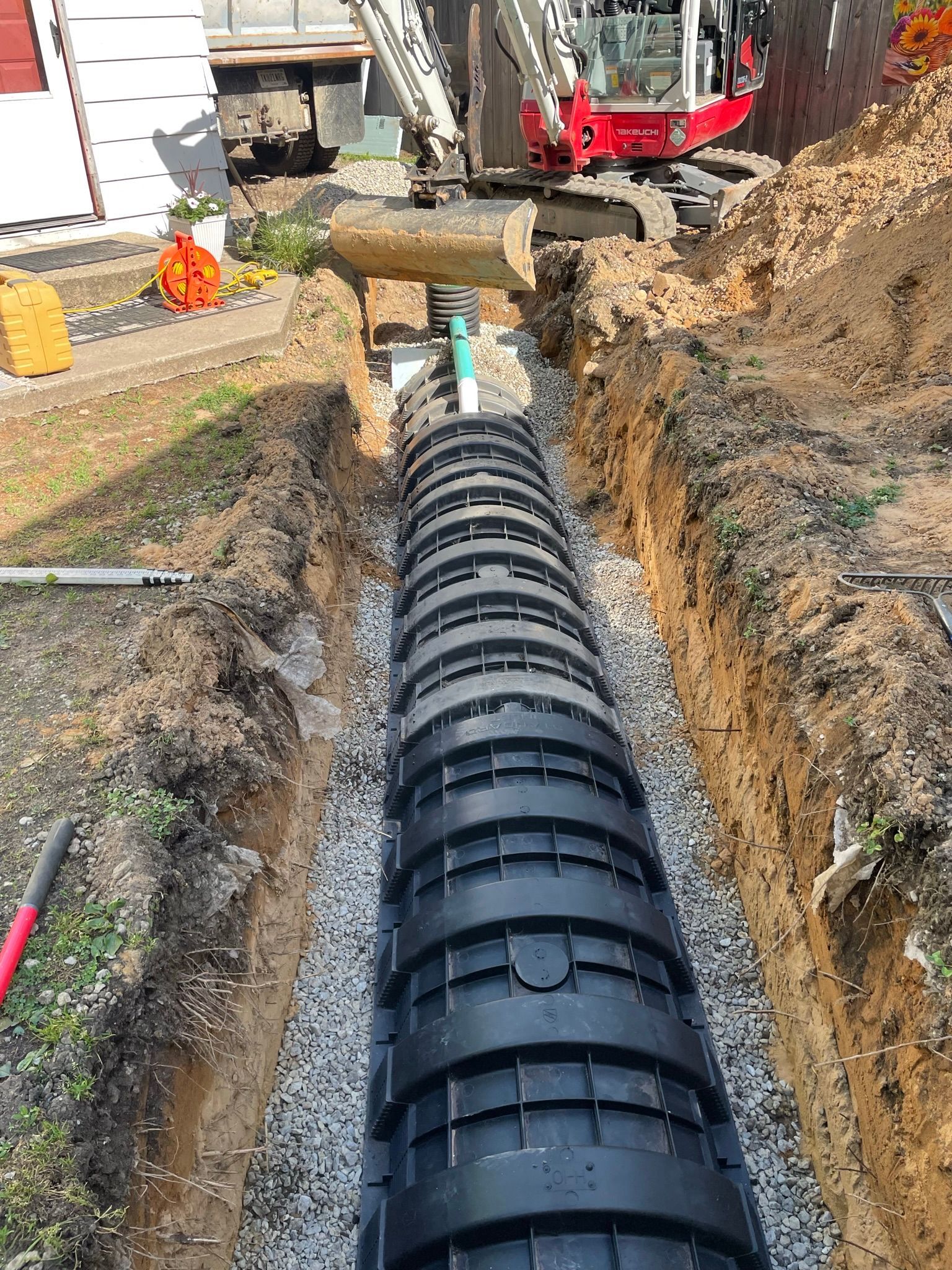 Construction site: long trench with black drainage system. An excavator is in the background.