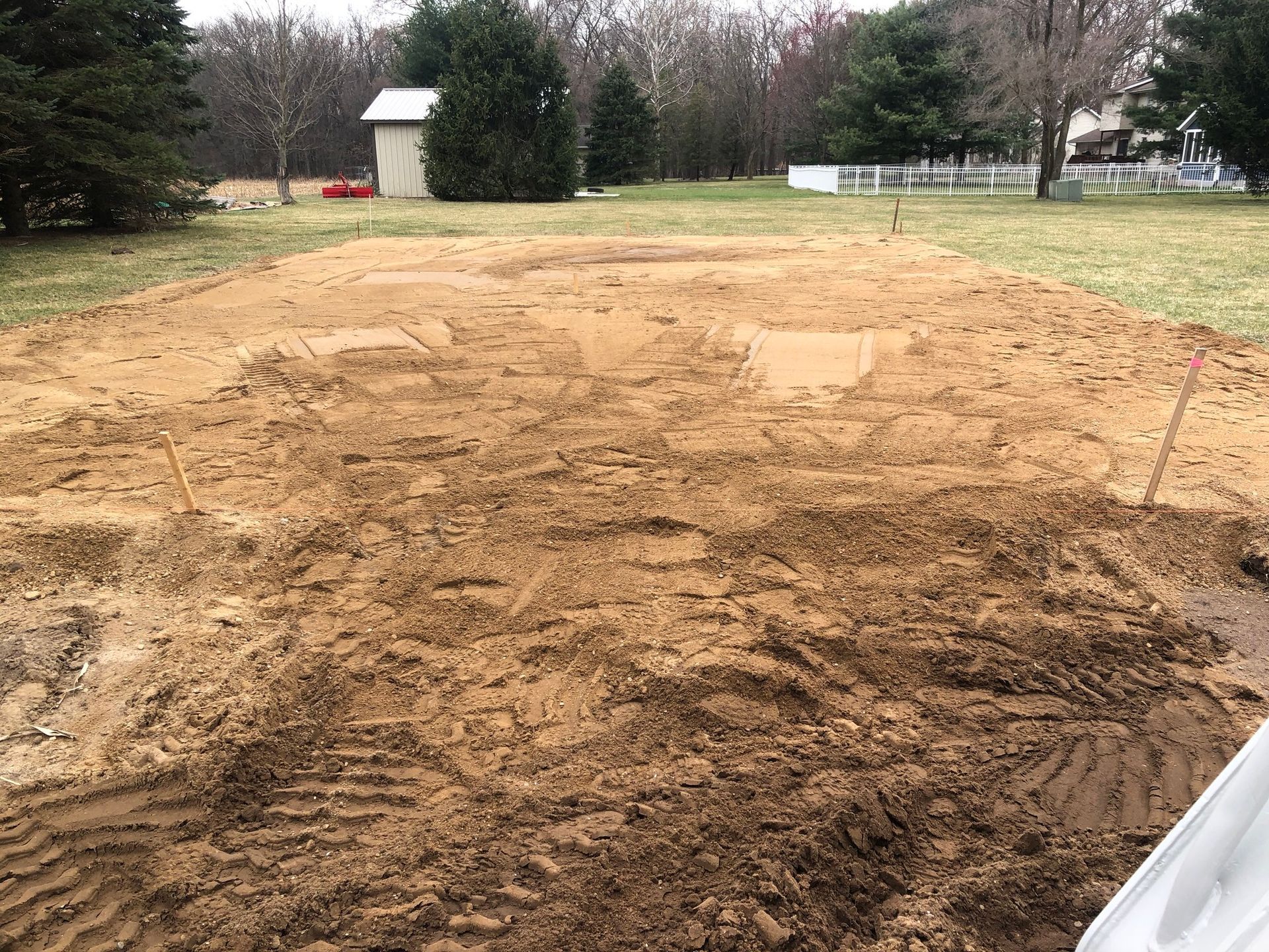 Excavated dirt area in a grassy yard, marked with stakes; small shed and trees in the background.