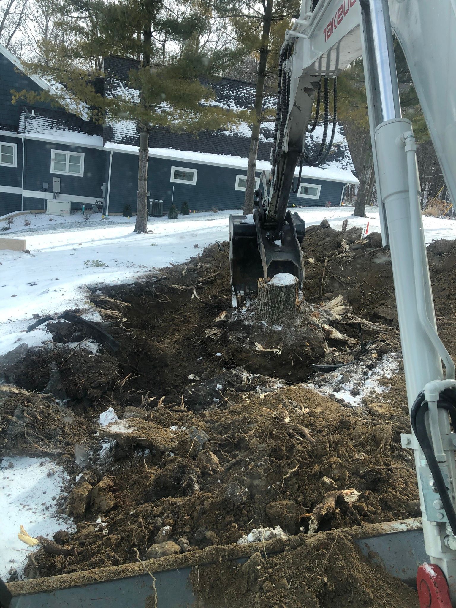 Excavator digging in dirt on a snowy landscape, residential building in the background.