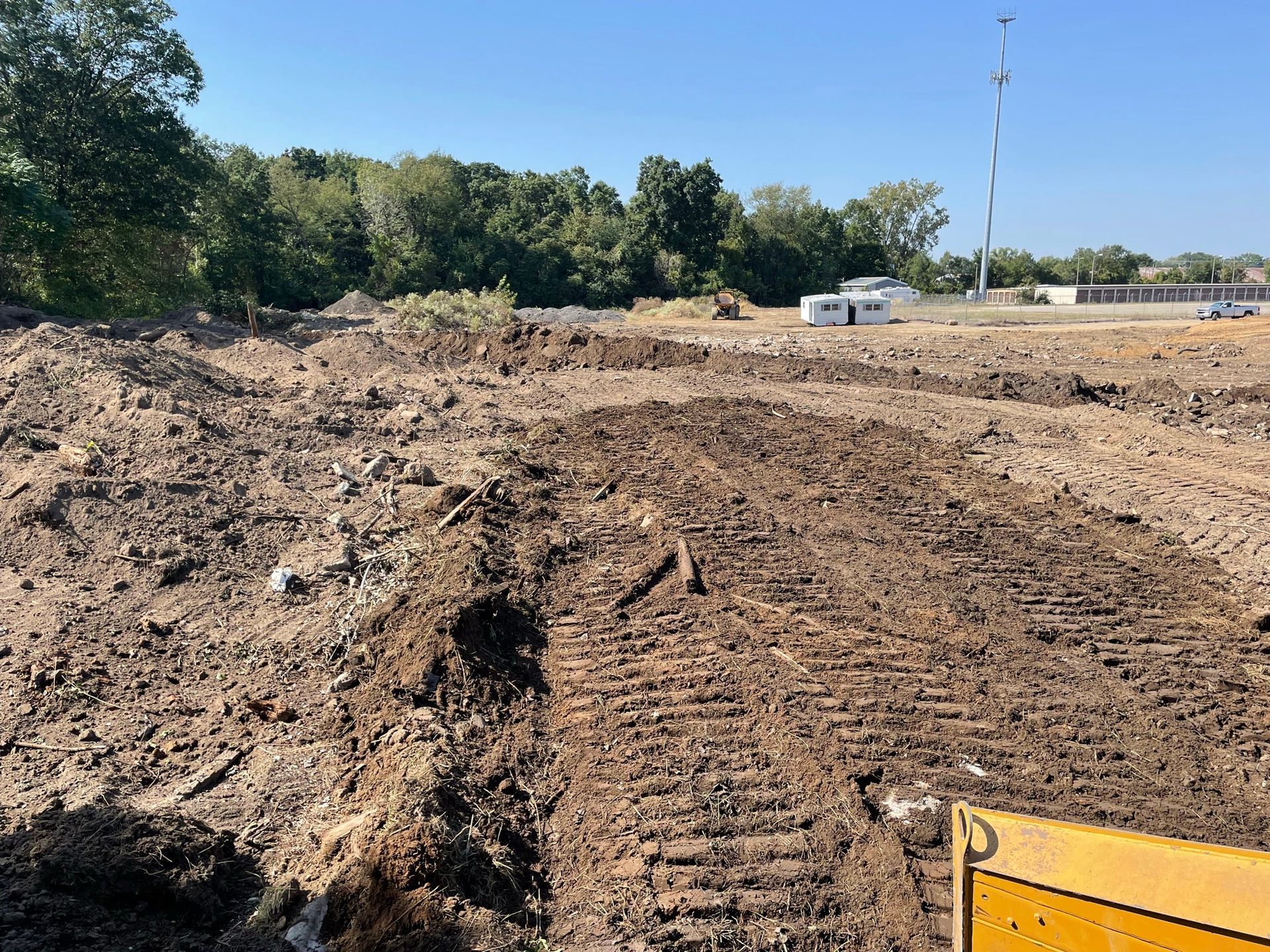 A construction site with disturbed soil, equipment, and a tree line under a clear blue sky.