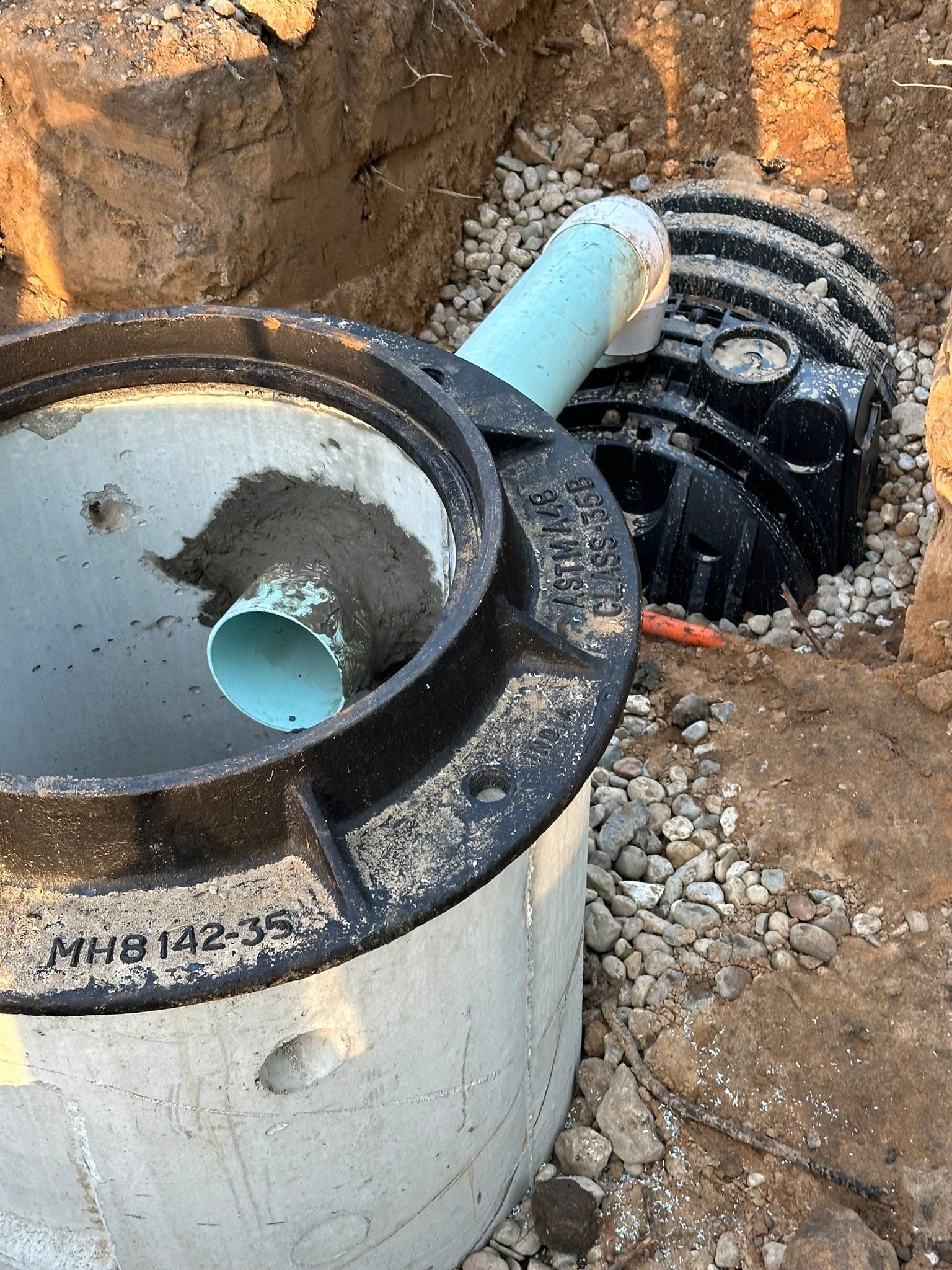 Concrete septic tank with connected pipes, partially buried in a gravel-lined trench.