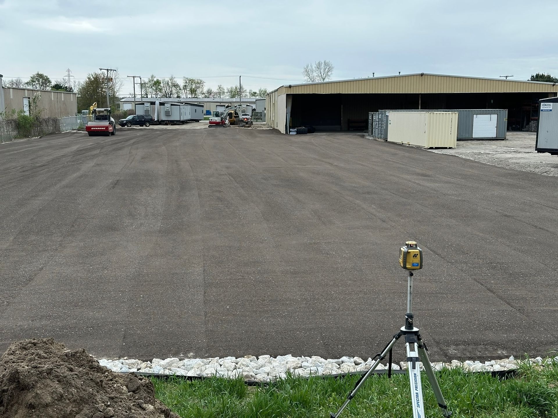 Asphalt paving of a construction site with a surveyor's instrument in the foreground and equipment working in the distance.