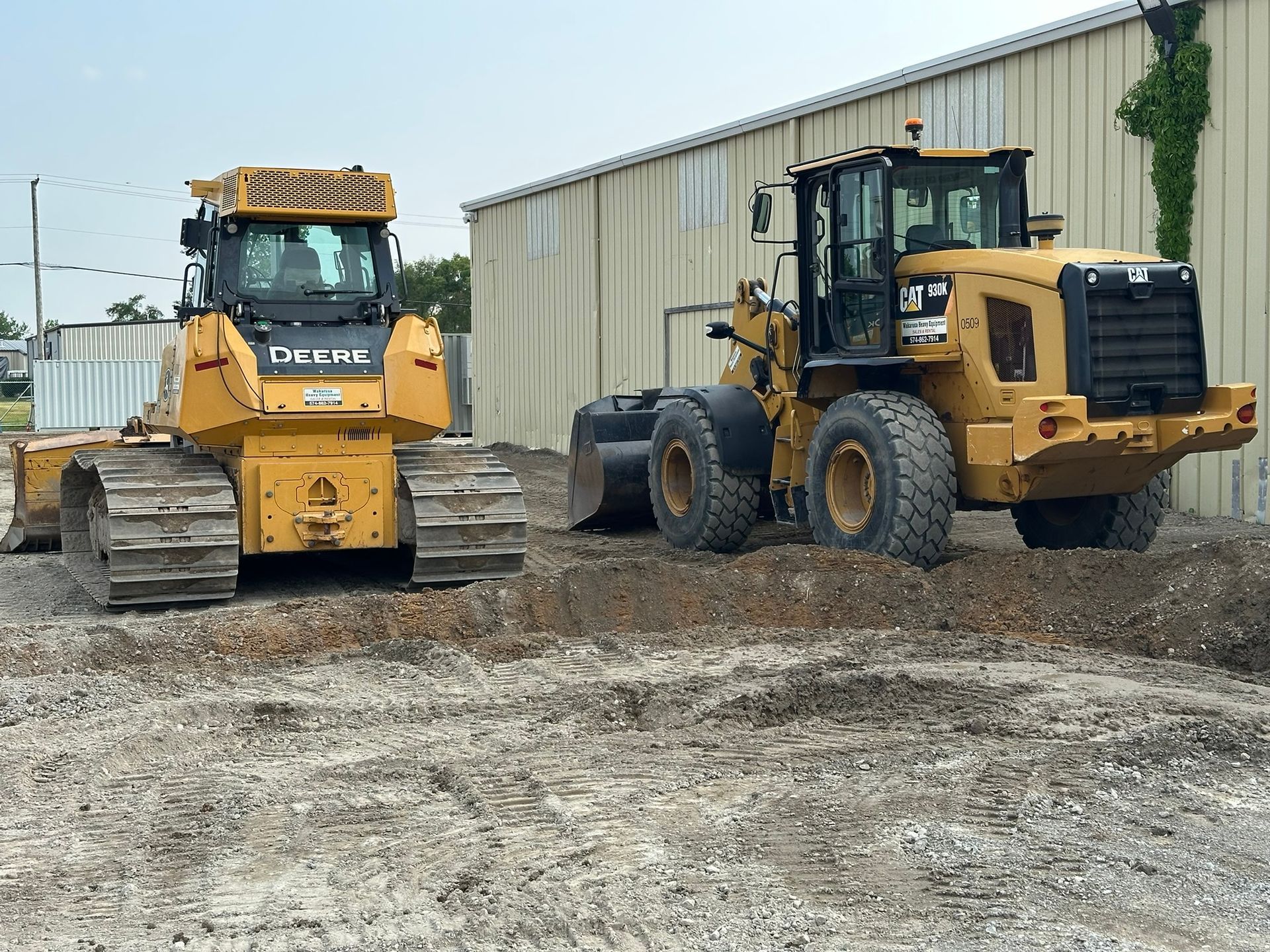 Two yellow construction vehicles on a gravel lot near a building.