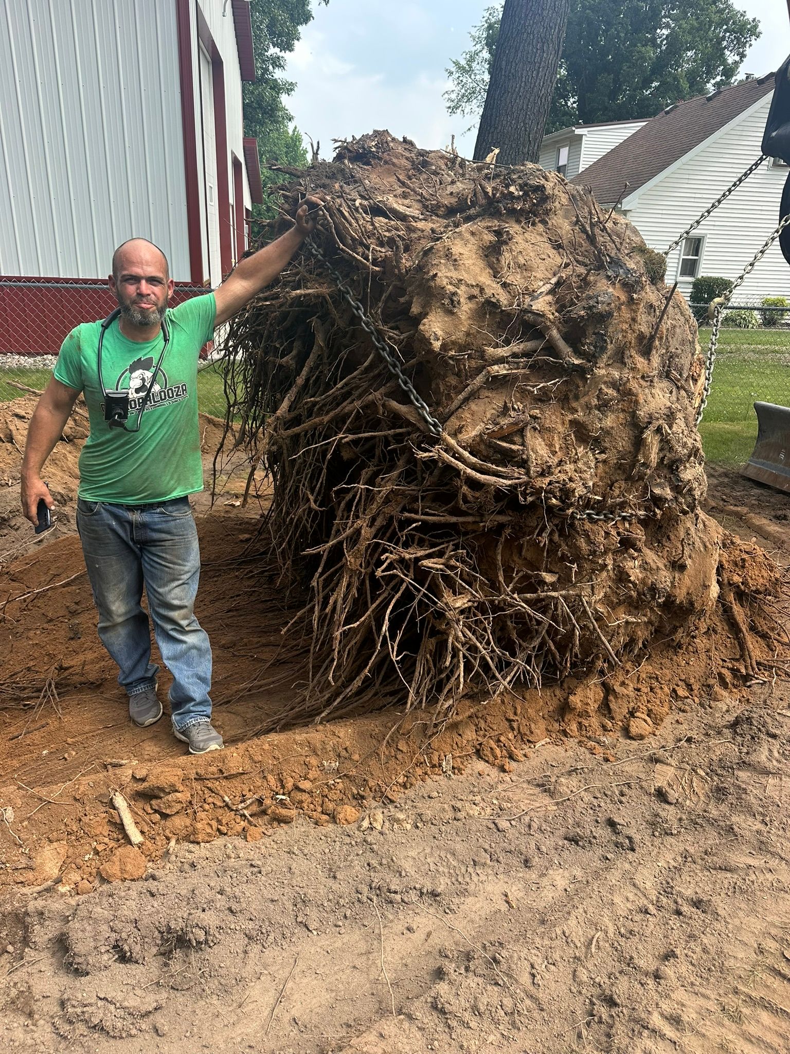 Man standing next to large tree root ball, red and white building in background.