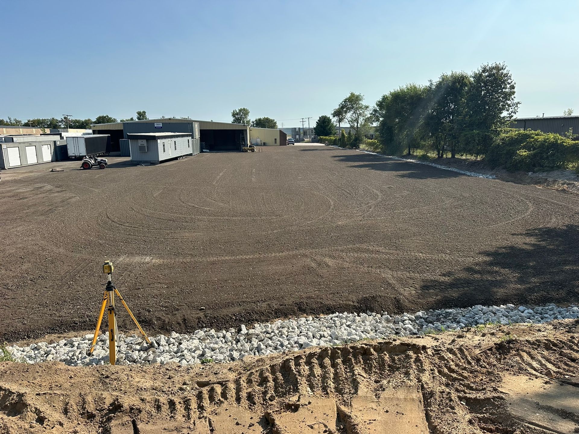 A leveled construction site with dirt and gravel, containers in the background, and a sunny sky.