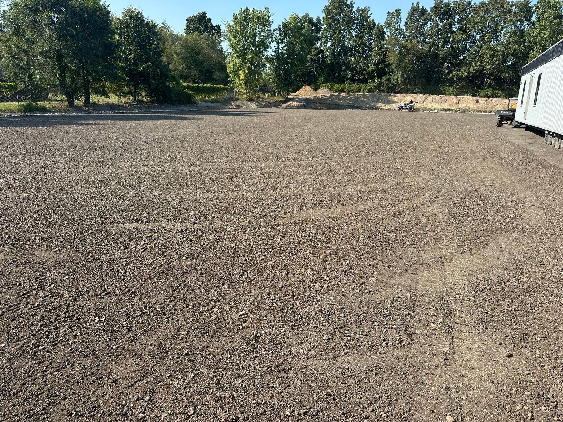 Gravel-covered lot with trees in the background under a clear sky. A structure is on the right side.