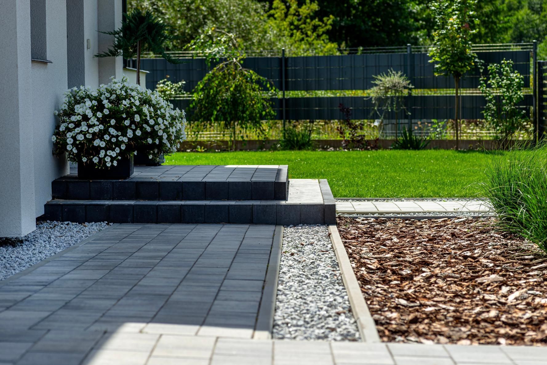 Modern home exterior with gray paved patio, steps, and flower pots; green lawn in the background.