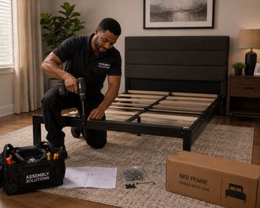 Man assembling a black metal bed frame in a bedroom with tools and parts on the floor.