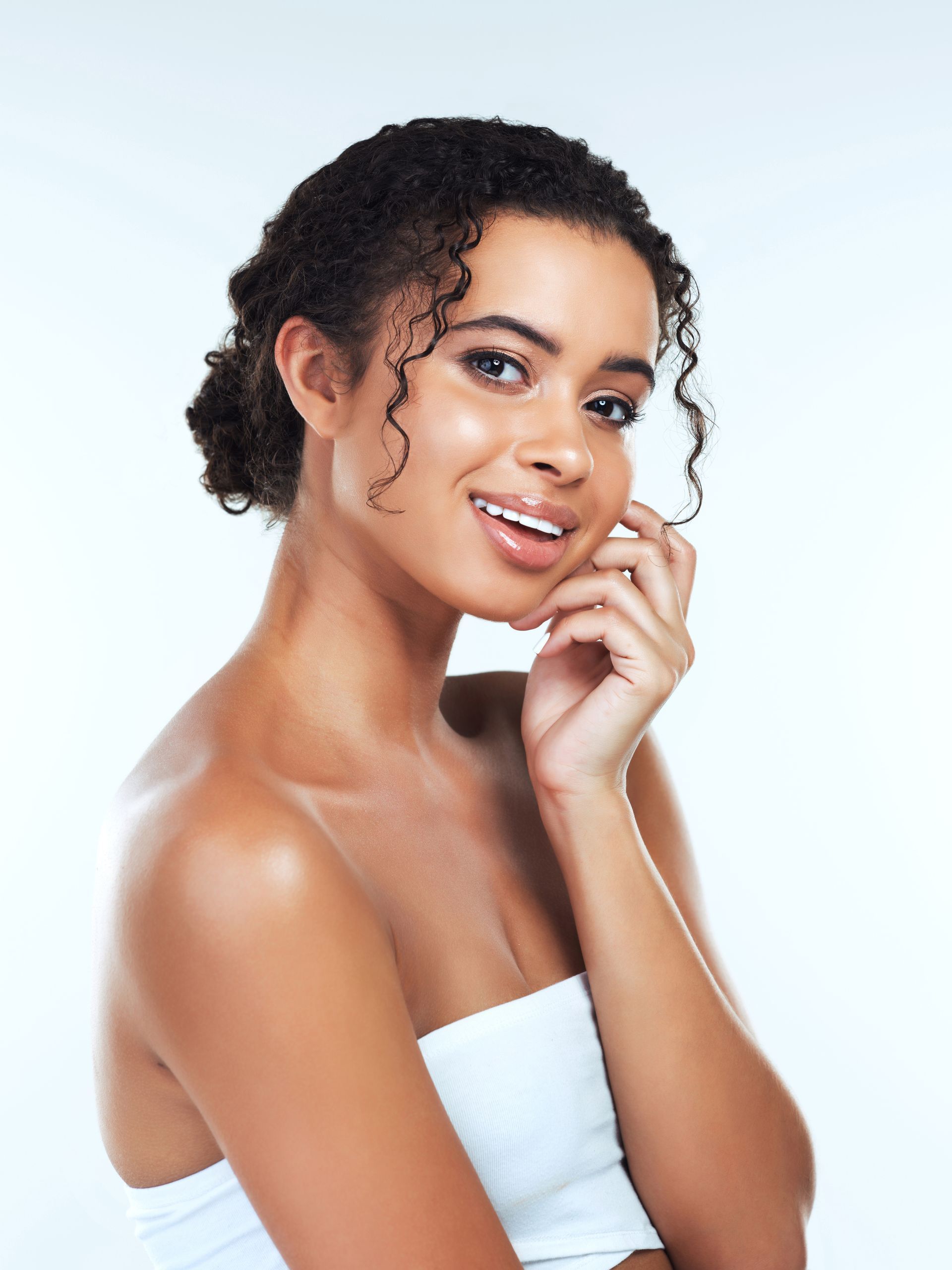 A woman with curly hair is smiling and touching her face.