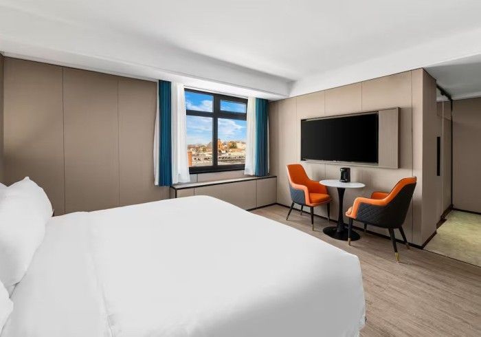 Hotel room with a bed, TV, seating area with orange chairs, and a window with a city view.