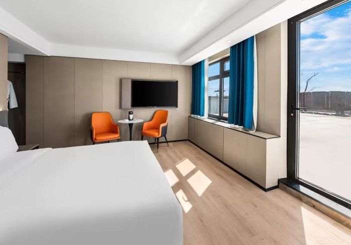 Hotel room with white bedding, orange chairs, and a large window overlooking a view.