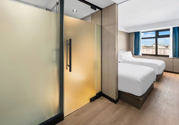 Hotel room with two twin beds, frosted glass shower door, and window with blue curtains.