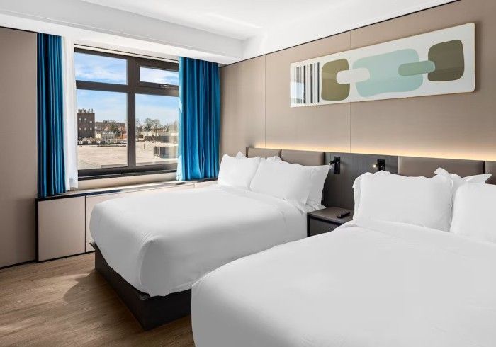 Hotel room with two queen beds, window with city view, blue curtains, and abstract wall art.