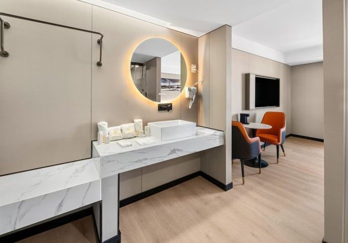 Modern hotel bathroom with a lit circular mirror, marble vanity, and seating area.