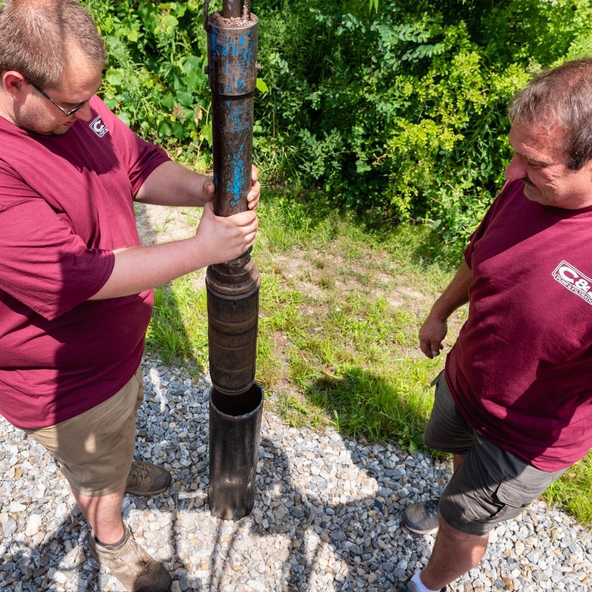 Two men in maroon shirts inspecting a rusted metal pipe outdoors on gravel near greenery.