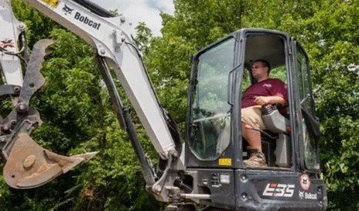 Operator in a Bobcat excavator cab clearing brush beside trees