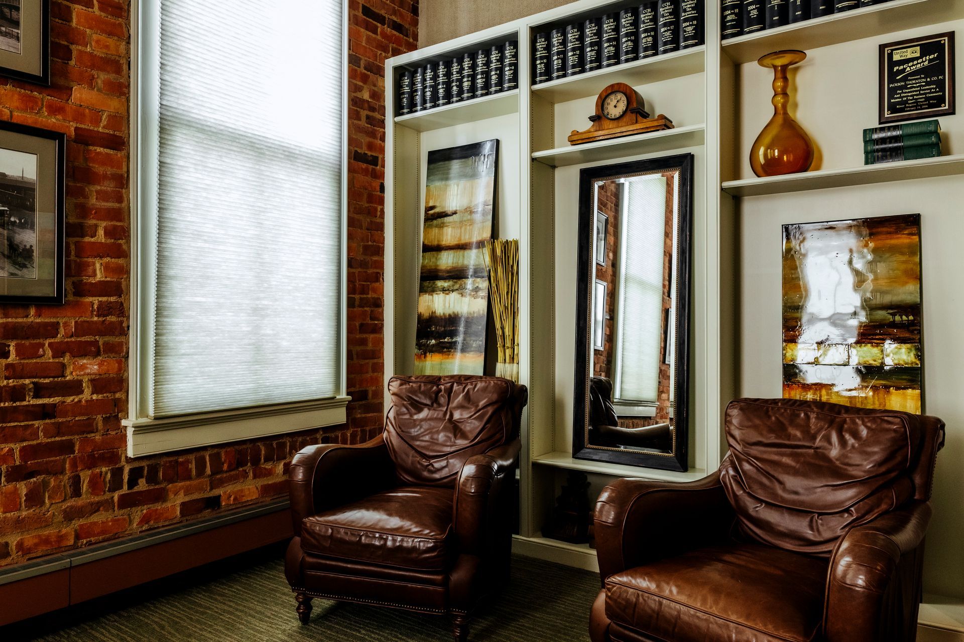 Two brown leather chairs in a cozy room with mirrors, framed art, and a brick wall