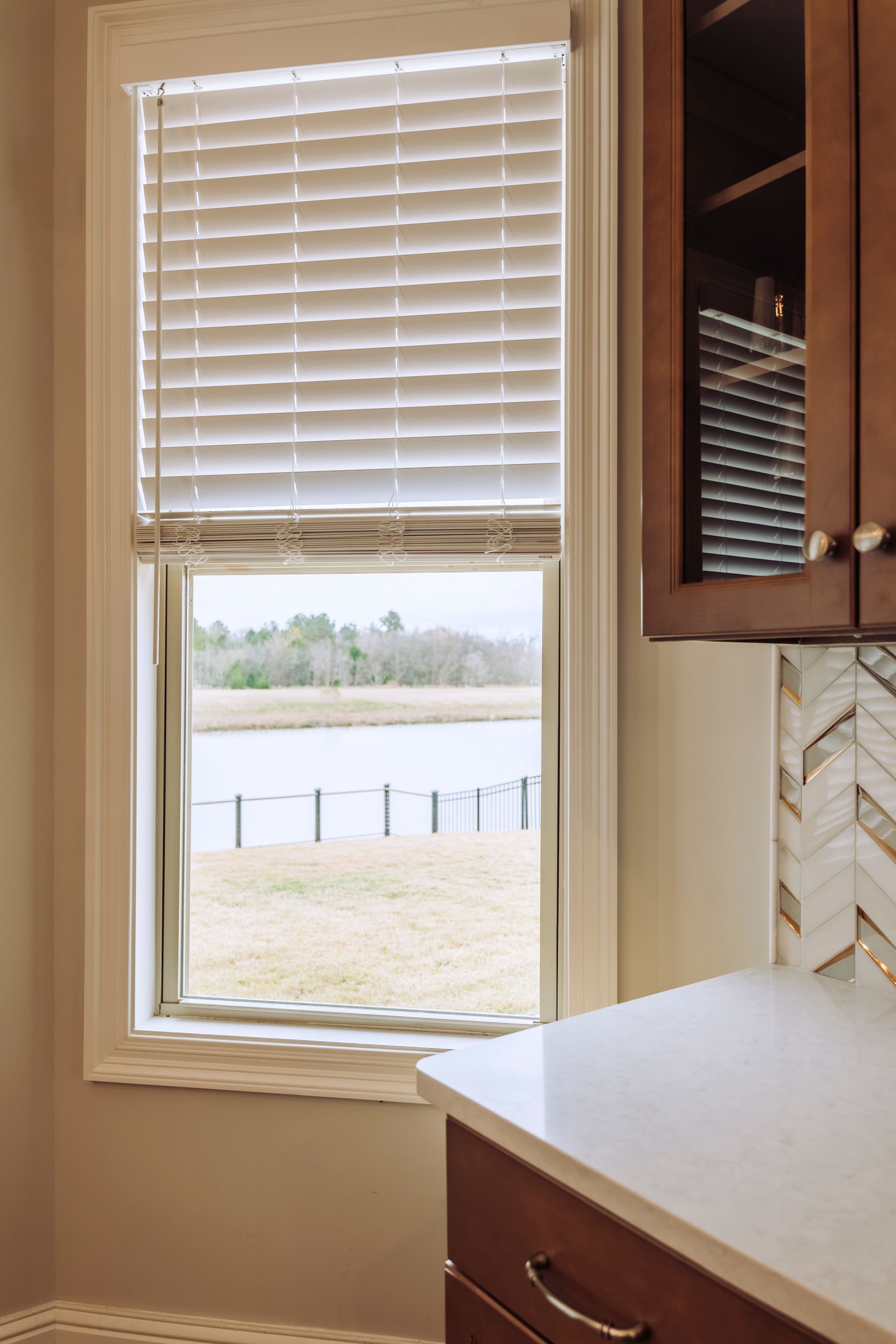 Kitchen window with white blinds, showing a lake view outside beside wood cabinets and a countertop