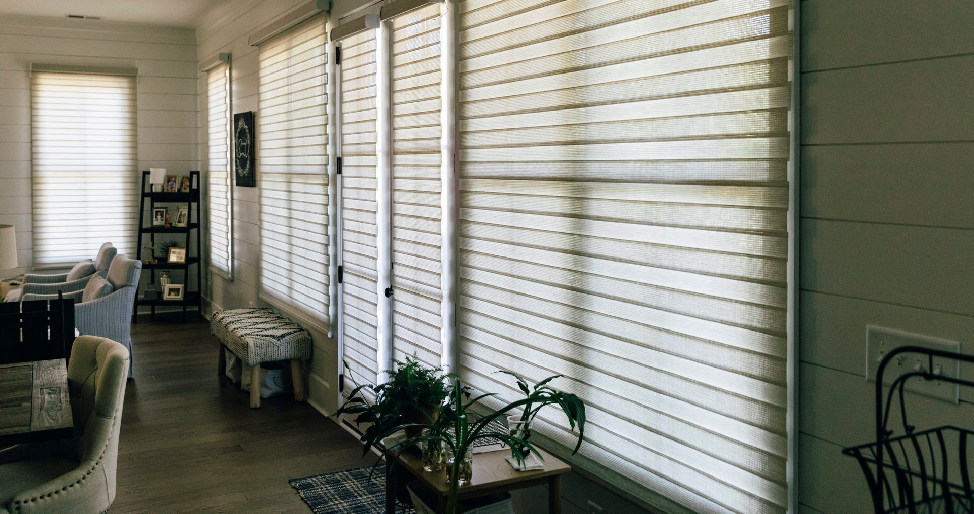 Bright porch with white blinds, potted plants, chairs, and natural light along the windows.