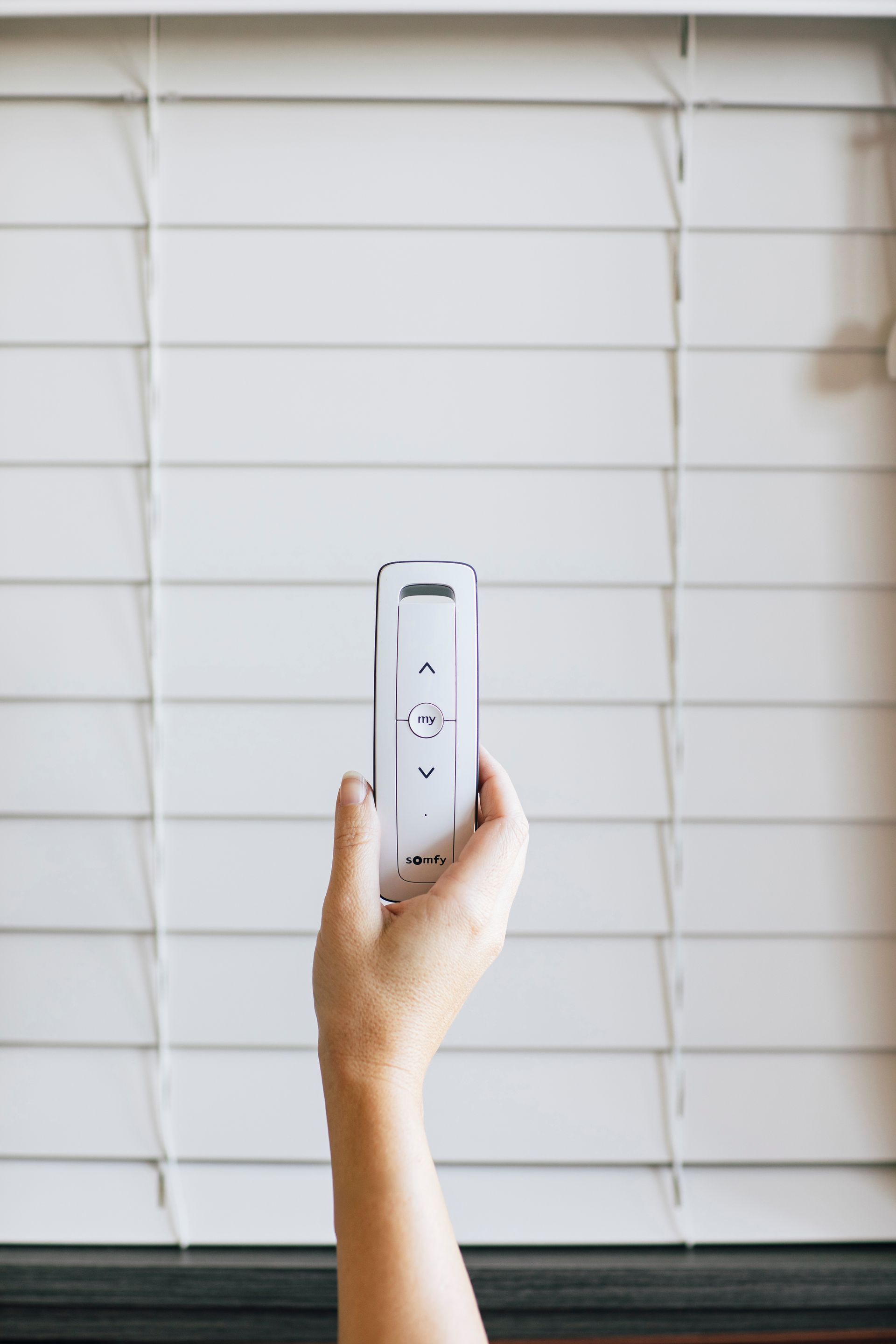 Hand holding a white remote in front of white window blinds