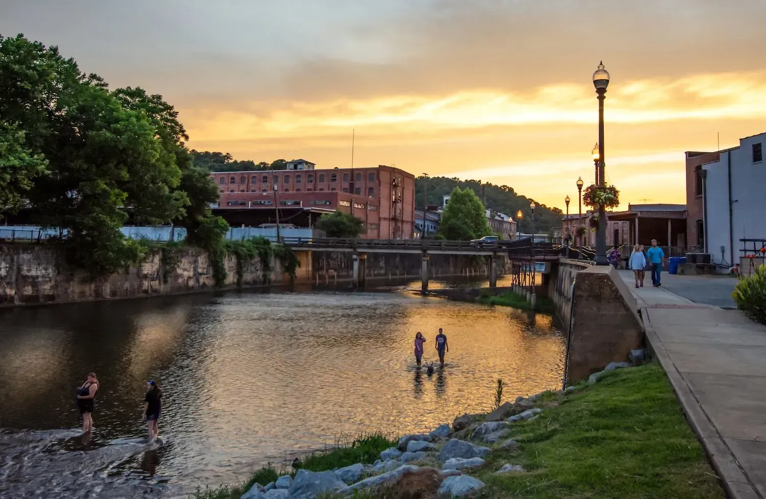 Sunset over a canal with people wading, a bridge, and riverside buildings and lamps