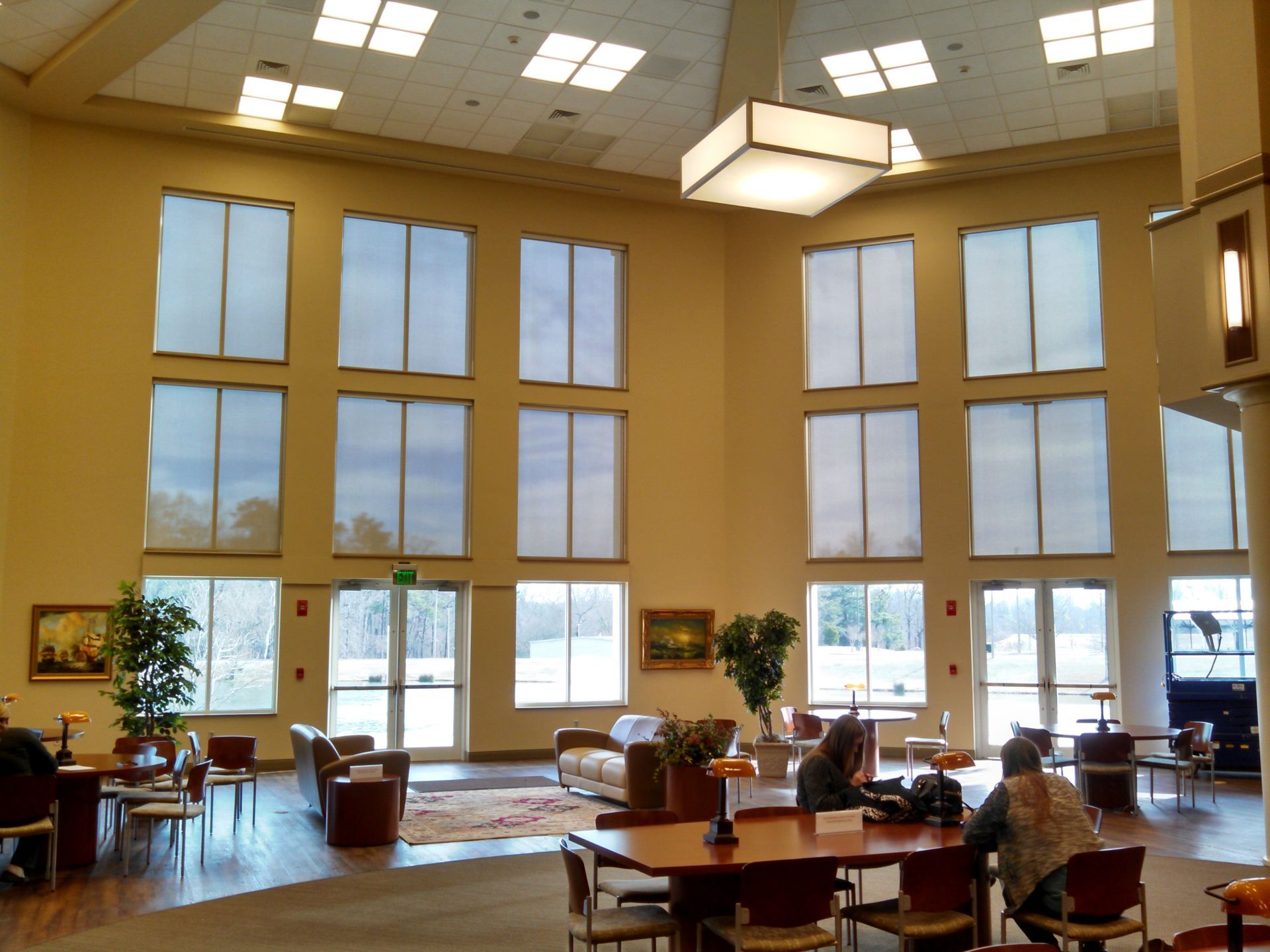 Bright lobby with tall windows, skylights, tables, chairs, and potted plants in warm daylight.