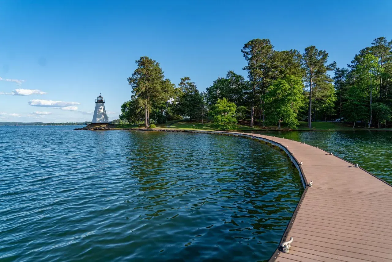 Curved lakeside boardwalk beside calm blue water, with trees and a distant lighthouse under a clear sky