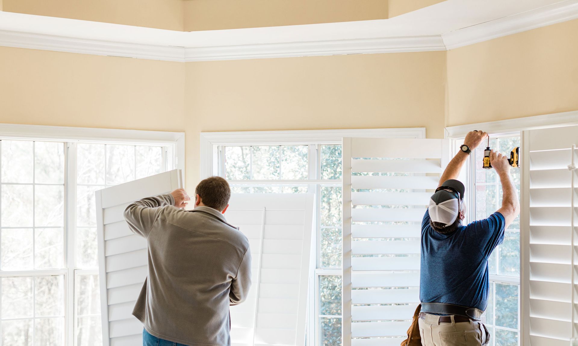 Two people installing white window shutters in a bright room.