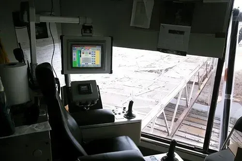 Interior cockpit view looking out over an airport apron and jet bridge, with a control screen and pilot seat in foreground