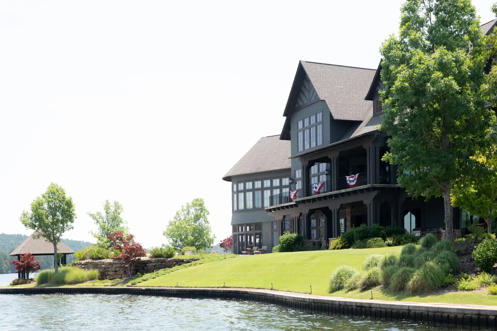 Lakeside manor with dark timber exterior beside a grassy bank and calm water under bright daylight