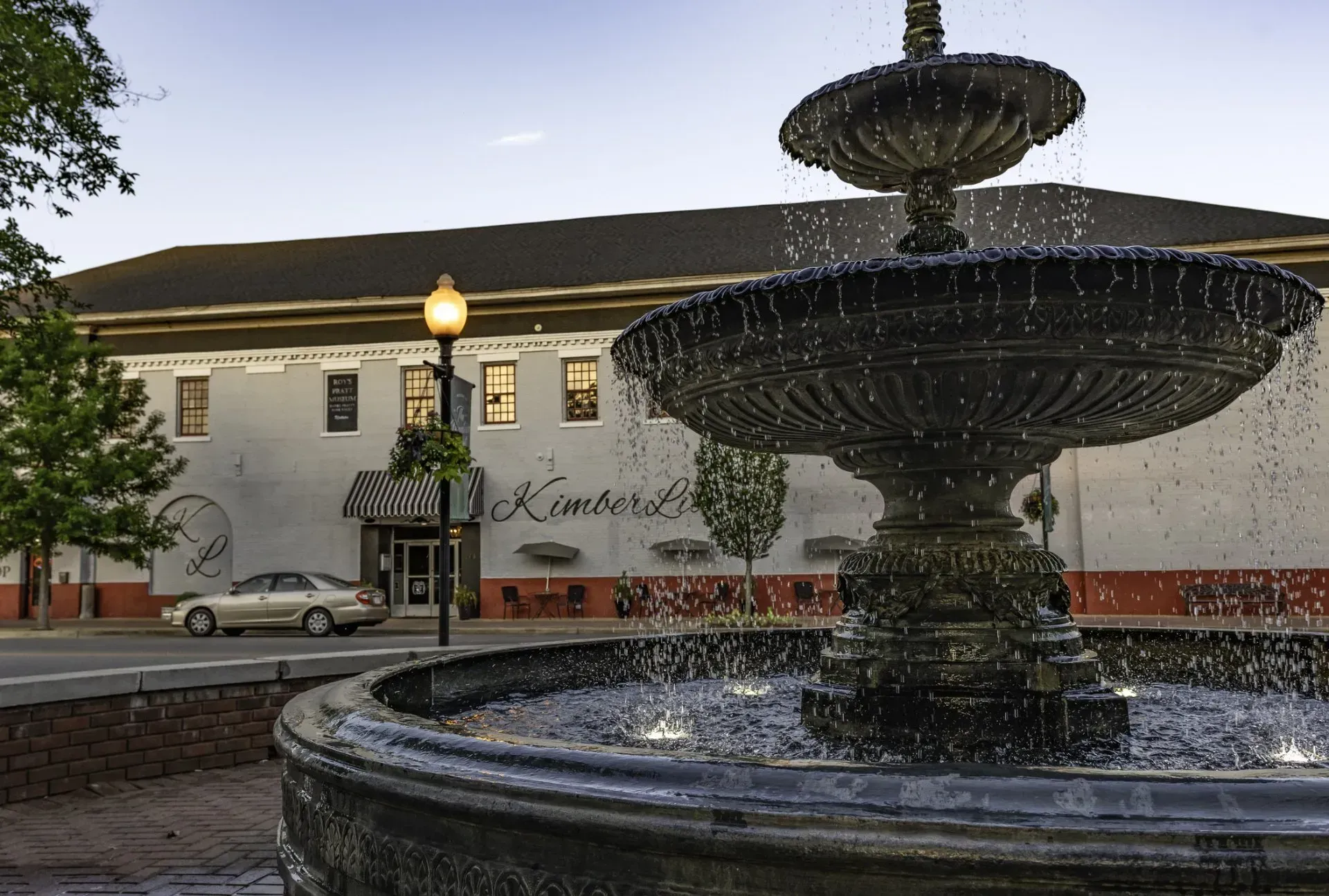 Fountain in front of a white building at dusk, with lit windows and flowers along the facade.