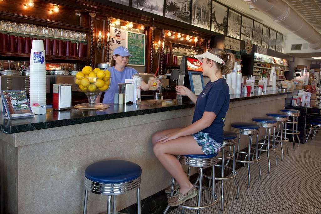 Two people at a diner counter with stools, one seated and one behind the bar near drinks and lemons