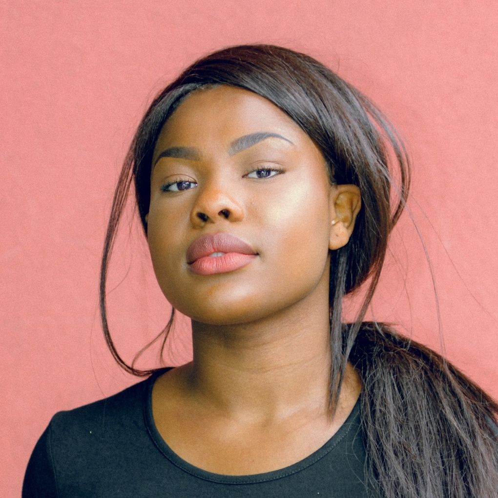 Young Black woman with long dark hair, looking directly at the camera, wearing a black shirt in front of a salmon-colored wall.