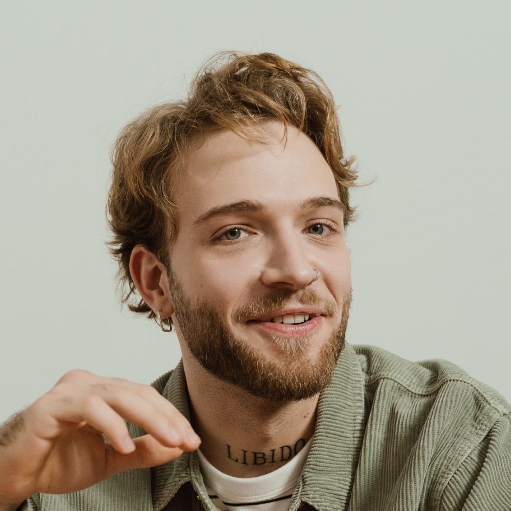 Smiling young man with light brown hair, wearing a green shirt, hand raised, and a 