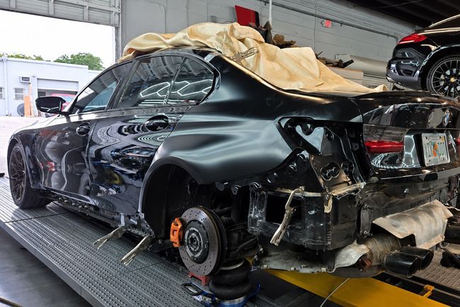 Damaged black sports car in a garage, rear end dismantled and wheel removed after a crash.