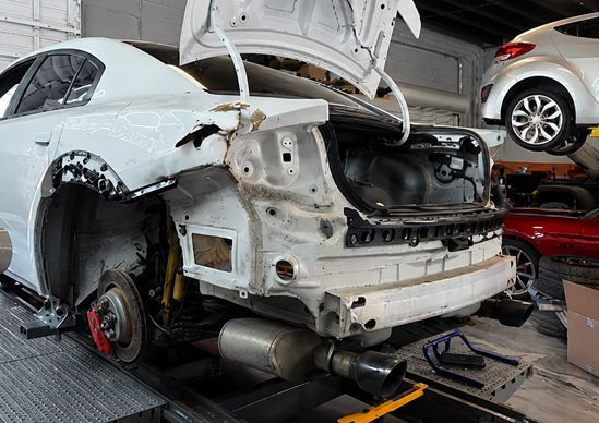 White car in a body shop with hood open, missing front bumper, and exposed rear wheel assembly under repair