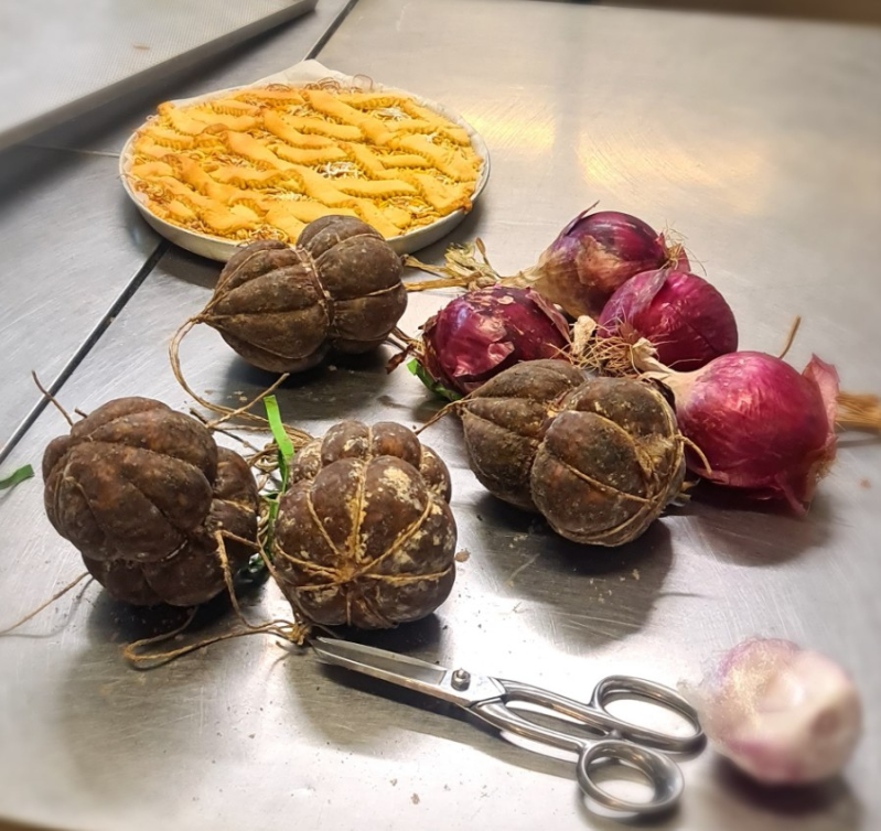 Sul bancone della cucina si trovano una torta rotonda, tre salsicce stagionate legate, diverse cipolle rosse, uno spicchio d'aglio e delle forbici.