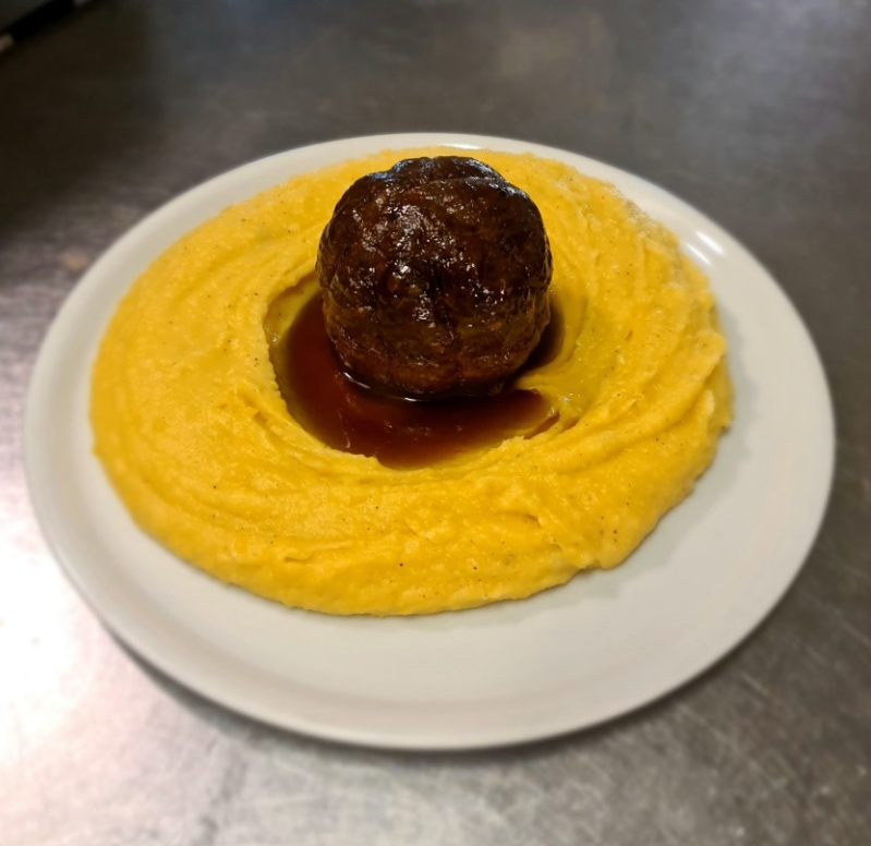 Una grande polpetta di colore marrone scuro, con al centro un anello di purè di patate cremoso e giallo, condita con una salsa marrone.