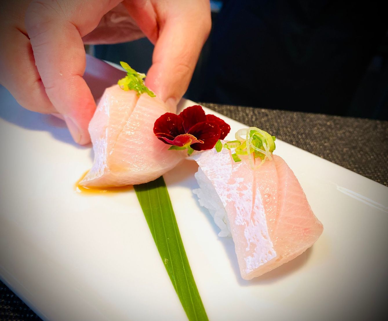 Hand placing two pink sushi pieces on a white plate, garnished with green herbs and red sauce.