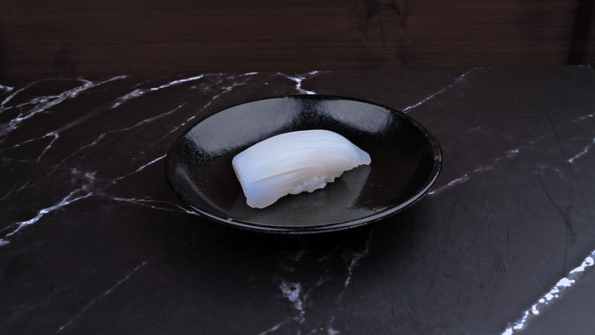White sushi-like piece on a black plate on a dark marble table.