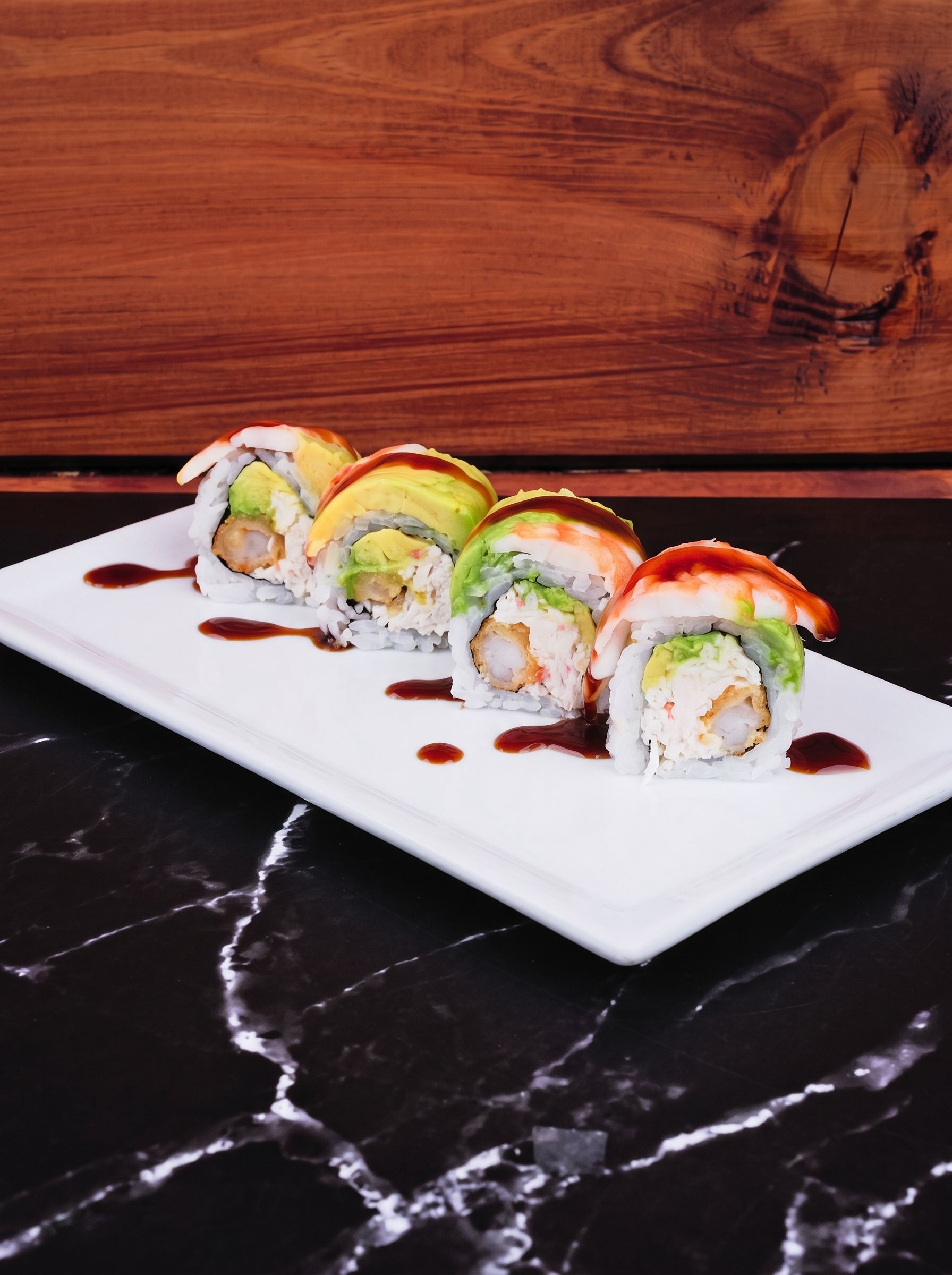 Sushi rolls on a white rectangular plate, topped with avocado, on a black marble table.