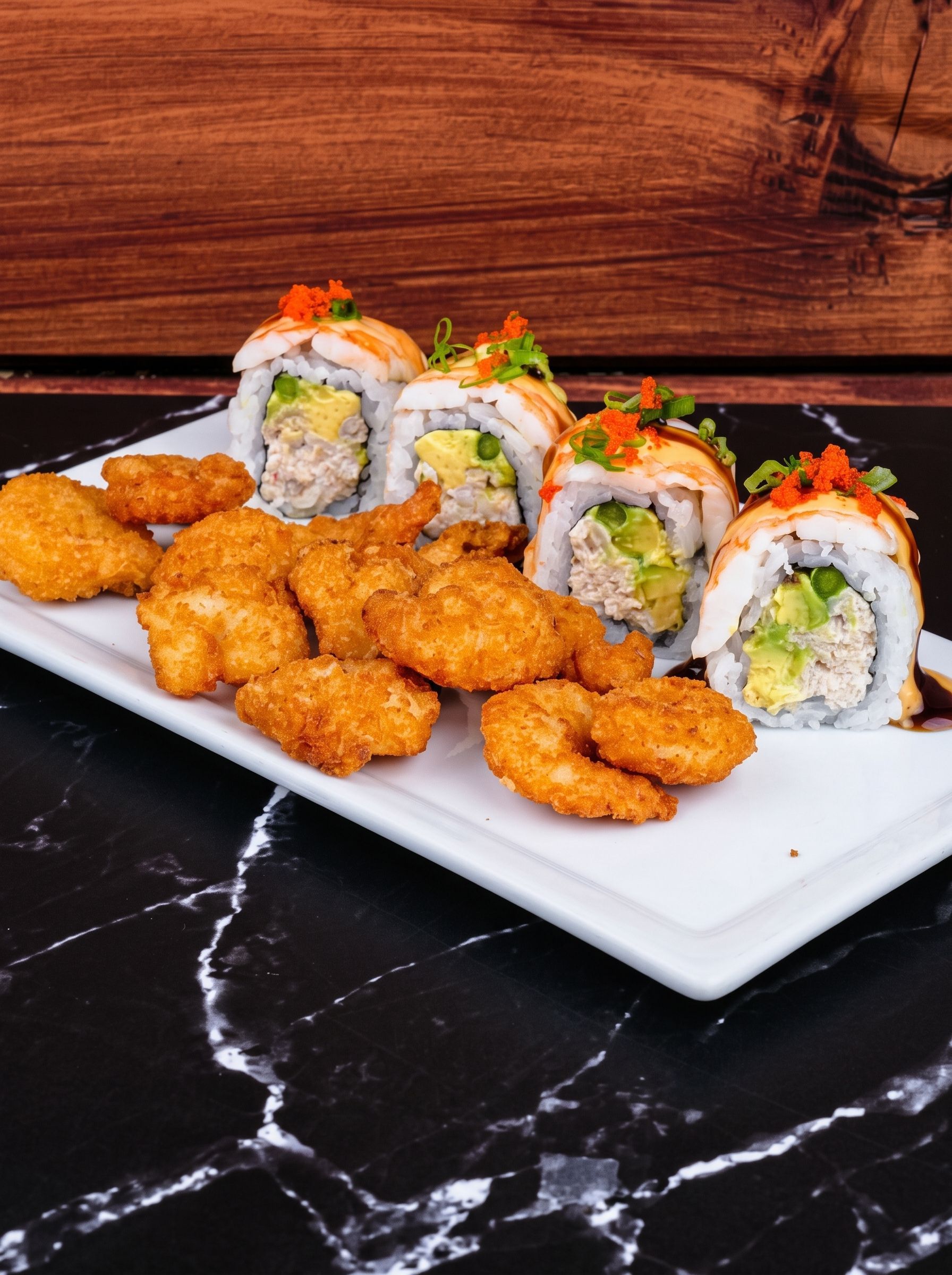 Sushi rolls with orange topping and crispy nuggets on a white plate against a dark marble table.
