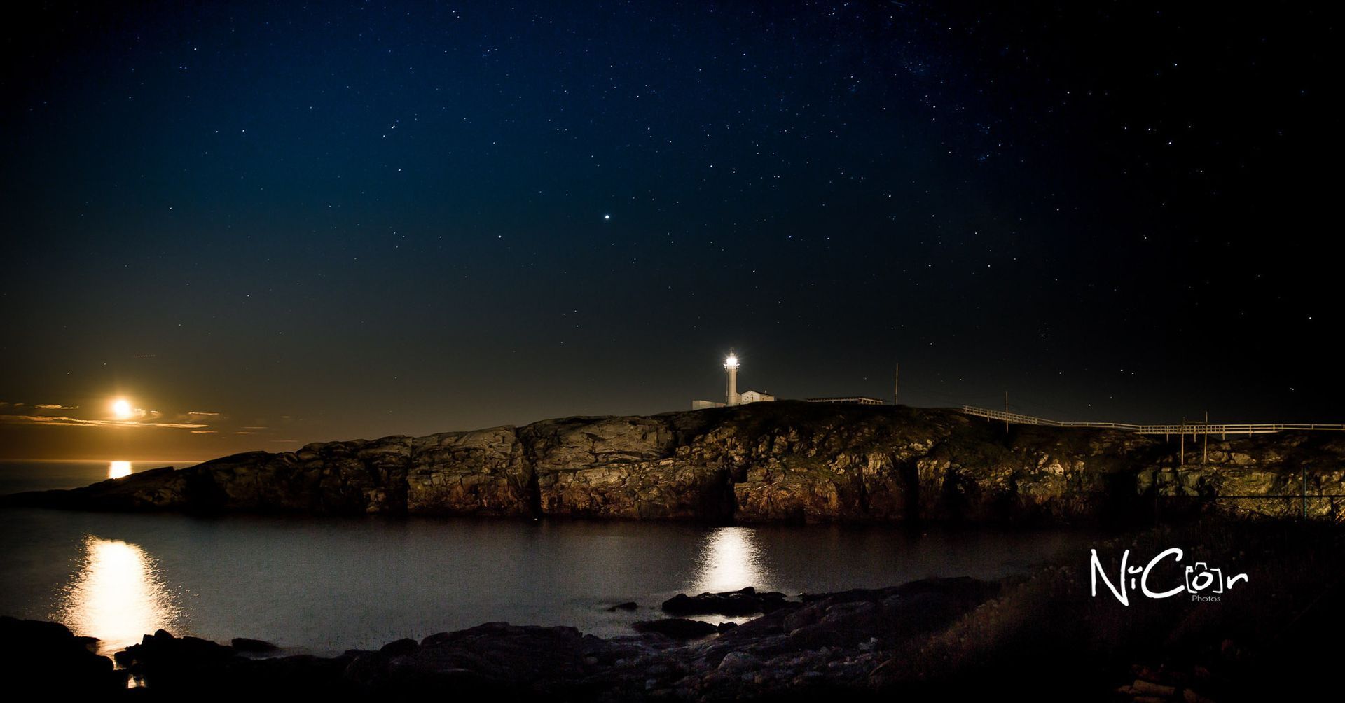 Night harbor with lighthouse on a rocky island, reflected on calm water under a starry sky.