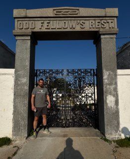 Stone gate labeled “Odd Fellow’s Rest” with a man standing beside it on a sunny day