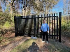 Person standing beside a black metal fence on a wooded path in daylight