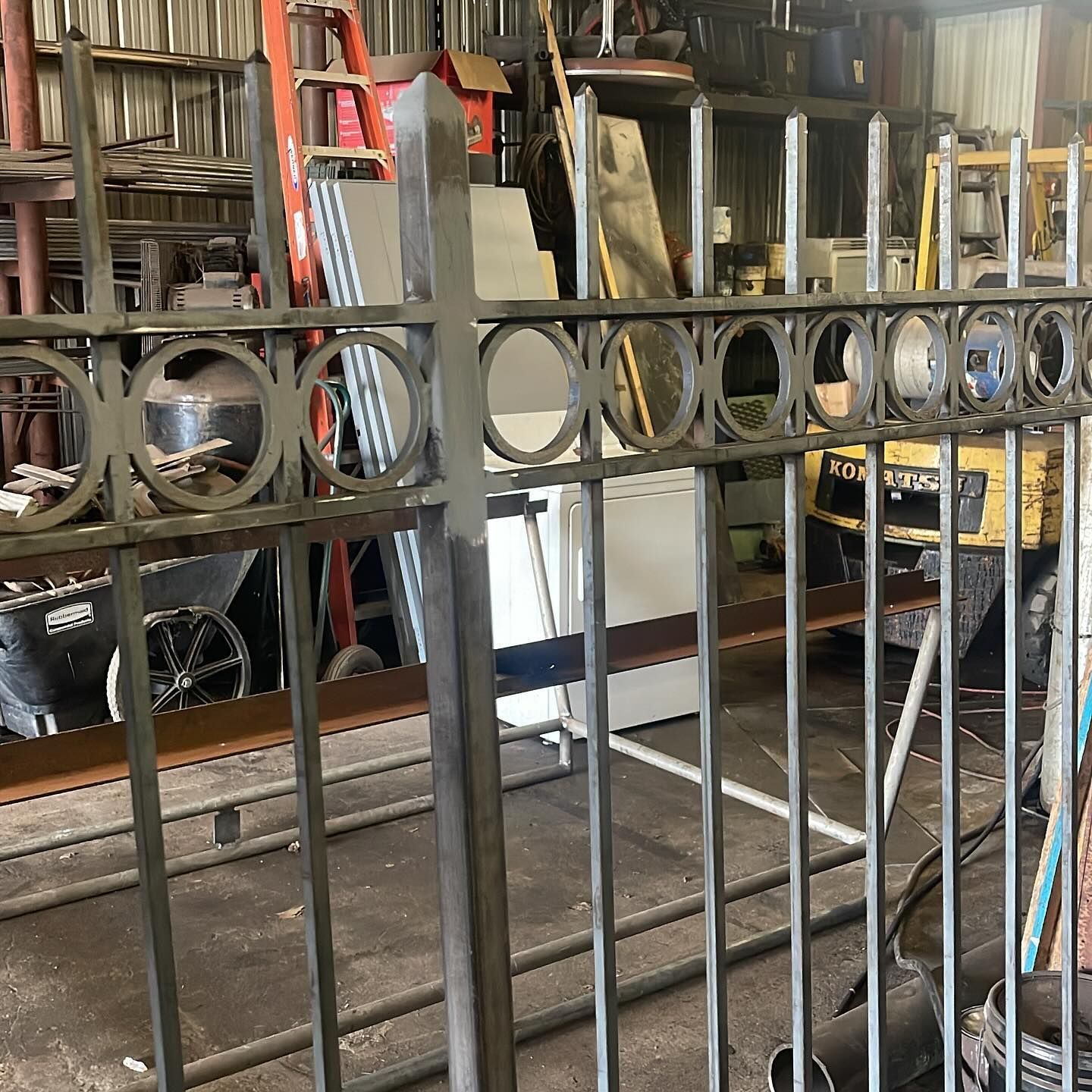 Metal fence with circular accents in a cluttered workshop, with a red ladder and shelving in the background