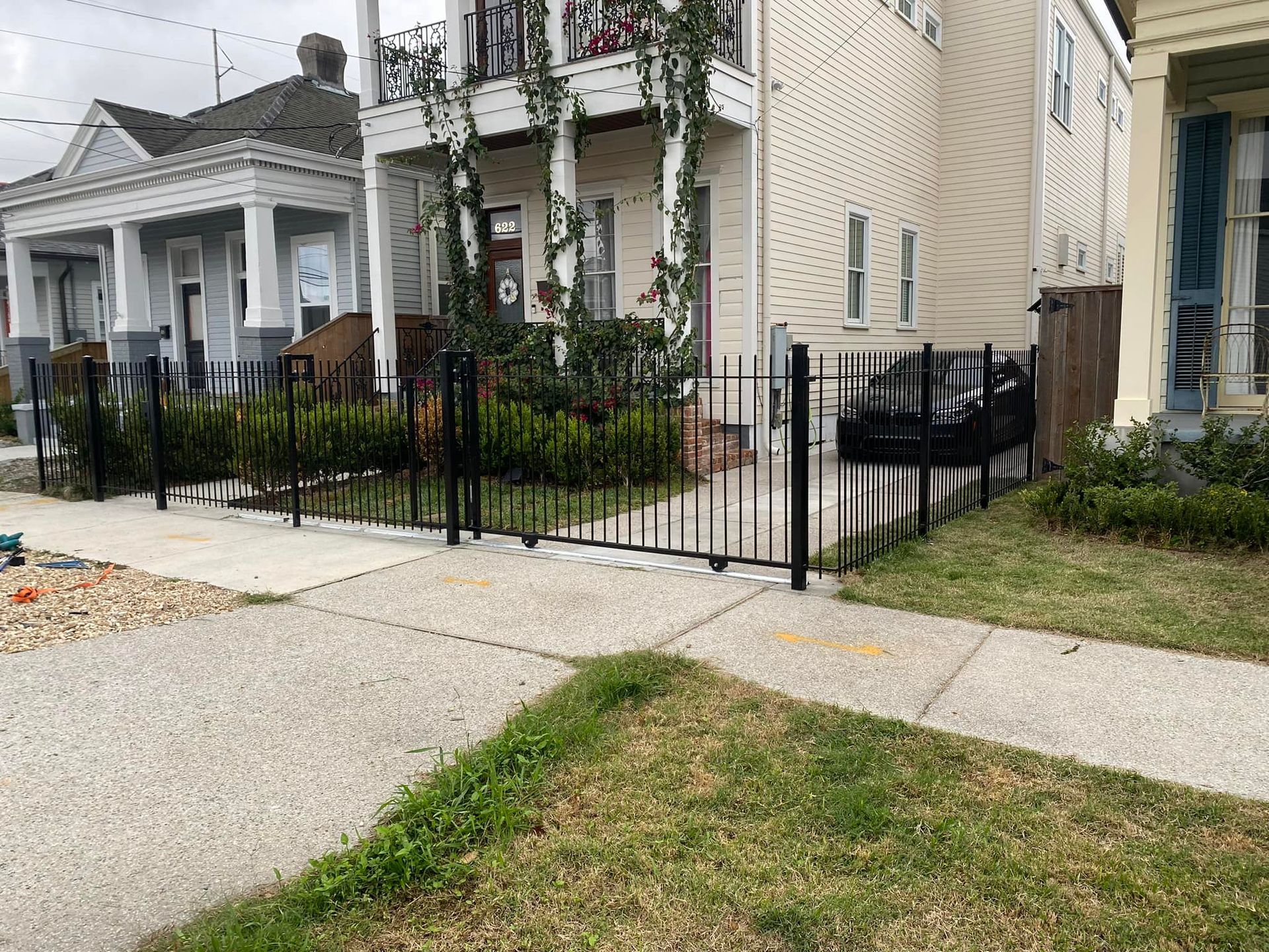 Black iron gate and fence beside a gray house along a sidewalk in a residential neighborhood