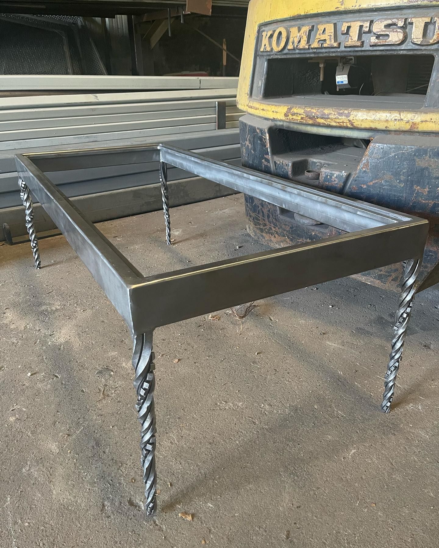 Metal frame table with twisted legs in a workshop, beside a Komatsu machine.